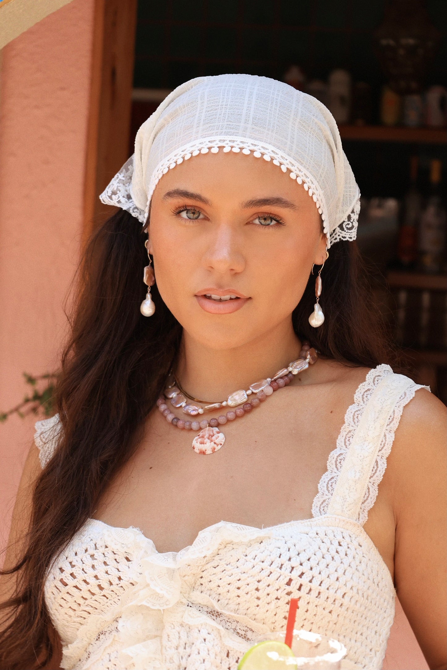 A woman wearing a white lace-trimmed headscarf and matching white crochet top, accessorized with layered pink and white necklaces and pearl drop earrings, set against a warm peach-colored wall background.