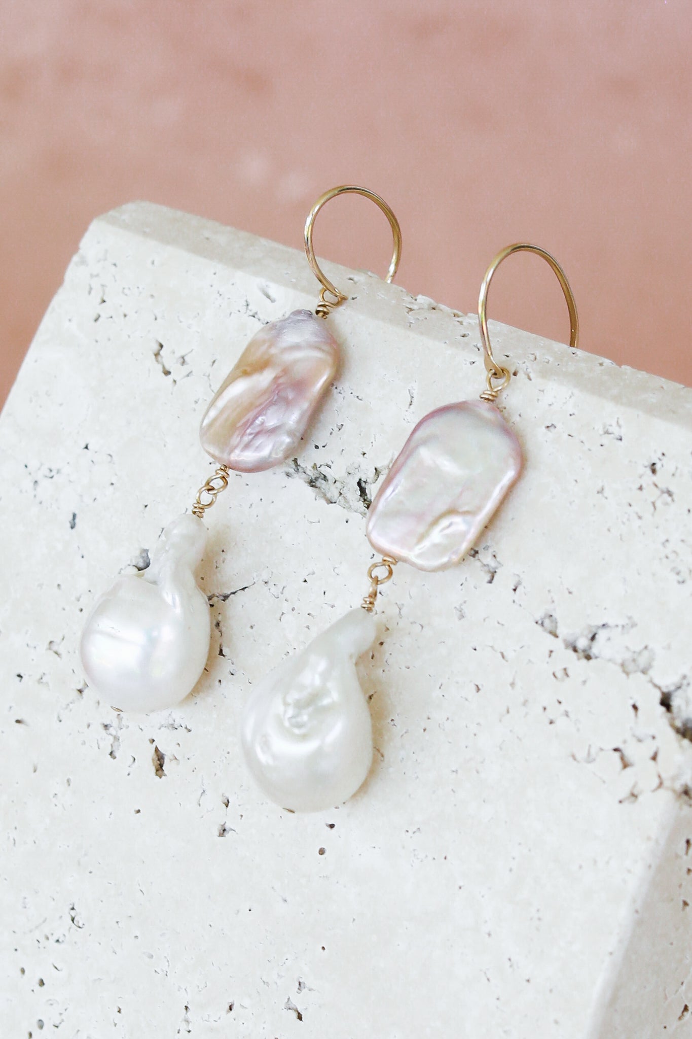 A pair of gold hoop drop earrings featuring irregular baroque pearls in soft pink and white tones, displayed on a textured white stone block against a dusty pink background.